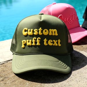 Puede incluir: Gorra de camionero verde oliva con el texto "Custom puff text" en letras amarillas en relieve. Otras gorras en rosa, negro y morado son visibles en el fondo. Las gorras están sobre una superficie de piedra cerca de una piscina.