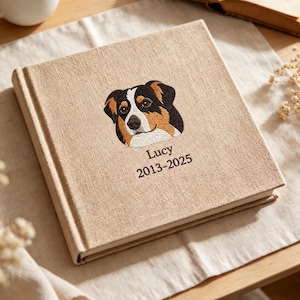 Könnte beinhalten: Ein quadratisches Fotoalbum mit beigem Leinenbezug und einer gestickten Darstellung eines dreifarbigen Hundes. Unter dem Hund sind der Name Lucy und die Jahreszahlen 2013-2025 in dunkelbraunem Garn auf den Einband gestickt.