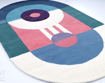May include: An oval-shaped rug with a geometric design. The rug features a combination of colors, including white, blue, pink, purple, and green. The design includes circles and rectangular shapes. The rug is made of a soft, textured material.
