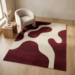 May include: A rectangular rug with a bold design. The rug features a deep red background with an abstract pattern in off-white. The rug is placed on a wooden floor, with a cream-colored armchair and a small wooden side table nearby.