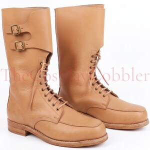 May include: A pair of tan leather boots with a lace-up design and a tall shaft. The boots feature a buckle closure on the side and a rounded toe. The boots are new and unworn.