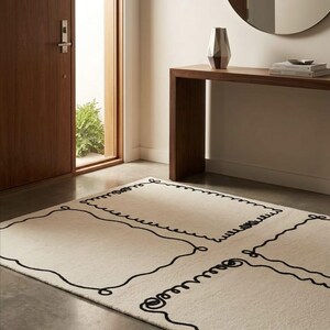 May include: A rectangular area rug featuring a cream-colored base with a bold, black, hand-drawn style line pattern that forms large, interconnected rectangular frames with wavy, scalloped edges across the entire surface of the floor covering.