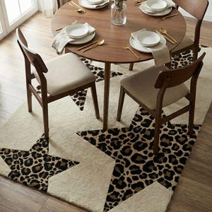 May include: A dining room scene featuring a round wooden table set for four with white plates, gold utensils, and beige napkins. Four wooden chairs with beige seats surround the table. A cream-colored rug with leopard print star designs is beneath the table.