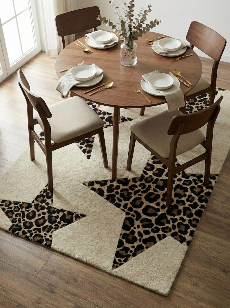 May include: A dining room scene featuring a round wooden table set for four with white plates, gold utensils, and beige napkins. Four wooden chairs with beige seats surround the table. A cream-colored rug with leopard print star designs is beneath the table.