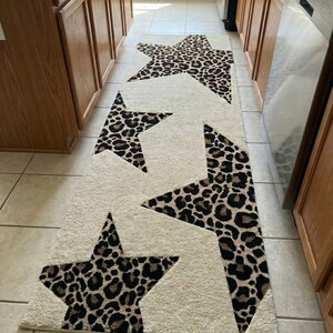 May include: Cream-colored runner rug with large leopard print star designs. The stars are in shades of brown and black, creating a bold contrast against the neutral background. The rug is placed in a kitchen or hallway.