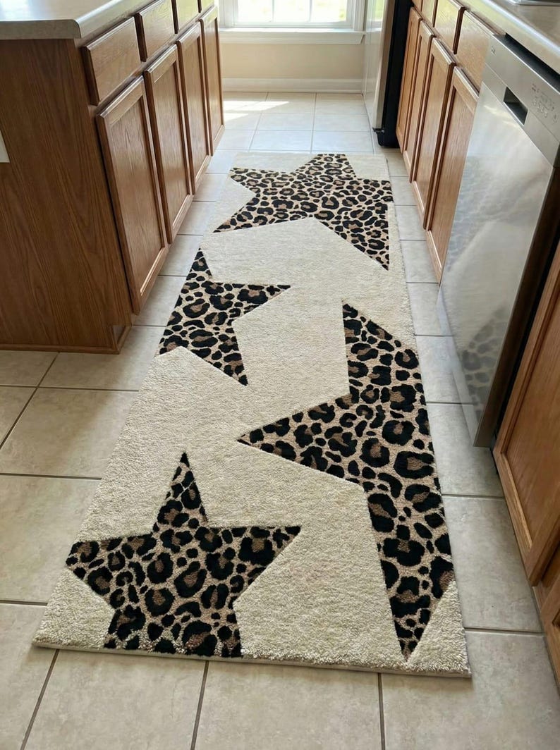 May include: Cream-colored runner rug with large leopard print star designs. The stars are in shades of brown and black, creating a bold contrast against the neutral background. The rug is placed in a kitchen or hallway.