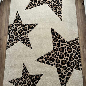 May include: A rectangular area rug with a cream-colored background and four large star shapes. The stars are filled with a brown and black leopard print pattern. The rug is made of a soft, plush material.