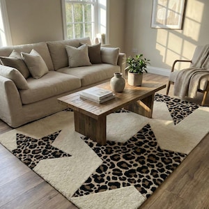 May include: A cream-colored area rug with large star designs featuring a leopard print pattern. The rug is placed in a living room with a beige sofa, wooden coffee table, and a neutral color scheme.