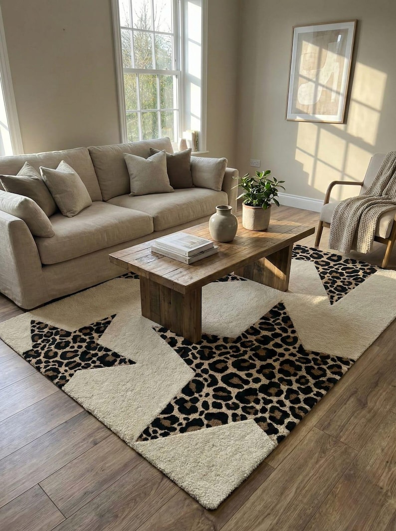 May include: A cream-colored area rug with large star designs featuring a leopard print pattern. The rug is placed in a living room with a beige sofa, wooden coffee table, and a neutral color scheme.