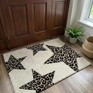 May include: Cream-colored rectangular rug with five large star shapes in a leopard print pattern. The rug is placed on a wooden floor in front of a brown wooden door. A fern plant in a pot and a woven basket are visible.