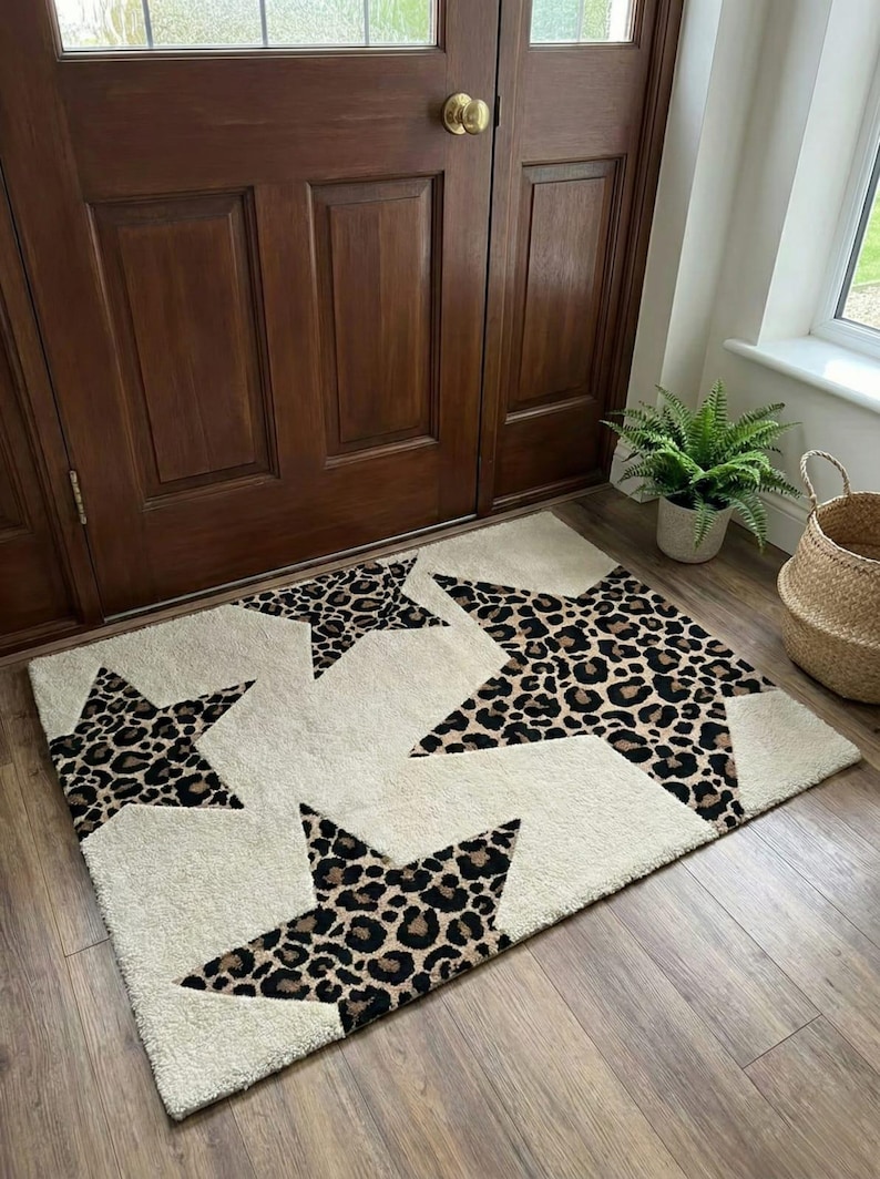 May include: Cream-colored rectangular rug with five large star shapes in a leopard print pattern. The rug is placed on a wooden floor in front of a brown wooden door. A fern plant in a pot and a woven basket are visible.