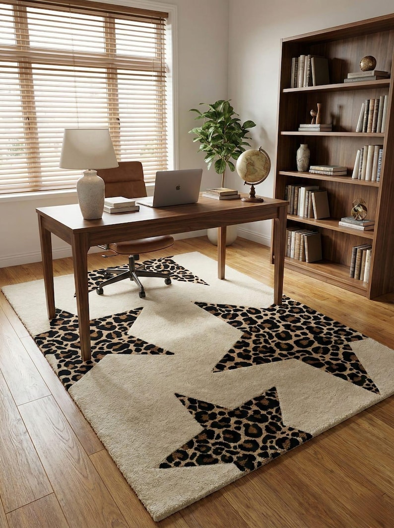 May include: A home office setup featuring a wooden desk with a laptop, lamp, and books. A cream-colored rug with large leopard print star designs sits beneath the desk and a rolling chair. A wooden bookshelf and globe are also visible.