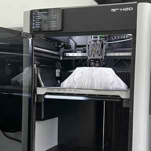 May include: A black and gray 3D printer with a clear door. The printer has a display screen and a printing bed with a white object inside. The printer is labeled "Bambu Lab H2D". The printing area is 350 x 320 x 325 mm.