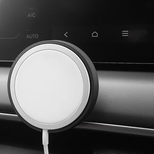 Puede incluir: Un cargador inalámbrico circular blanco con un borde plateado y un anillo exterior negro. Un cable blanco se extiende desde el cargador. El cargador está montado en un salpicadero negro, con controles de A/C y AUTO visibles.