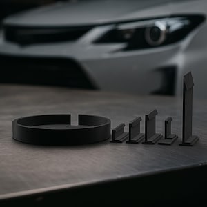 Puede incluir: Un conjunto de piezas automotrices negras, que incluye una base circular con una ranura y varias piezas rectangulares de diferentes alturas. Las piezas están dispuestas sobre una superficie oscura, con un coche en el fondo.