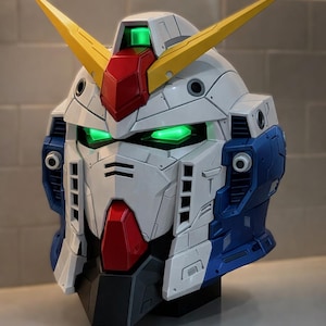 May include: A detailed model of a Gundam robot head, with a white, blue, red, and yellow colour scheme. The eyes and a top section glow green. The model is a detailed representation of the iconic mecha.