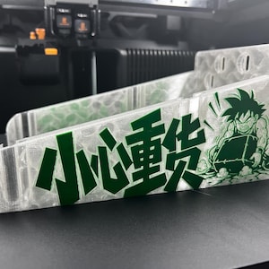 Puede incluir: Un contenedor de almacenamiento transparente y en ángulo con un gráfico verde de un personaje de dibujos animados y texto chino. El contenedor tiene ranuras y un diseño curvo. El texto dice "Manipular con cuidado".