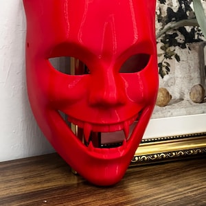 May include: A vibrant red mask with a devilish design. The mask features pointed horns, large eye openings, and a wide, toothy grin. The mask is made of a smooth material and is likely for costume or decorative purposes.