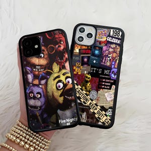 May include: Two black phone cases with colorful graphics. One case features characters from "Five Nights at Freddy's" with the text "Five Nights at" at the bottom. The other case has a collage design with the text "IT'S ME" and "CAUTION! KEEP OUT!"