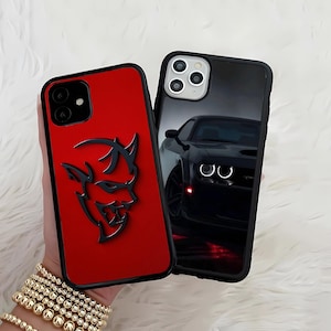 May include: Two phone cases. One is red with a black devil face design. The other is black with a car image. Both cases have a black border and are for a smartphone.