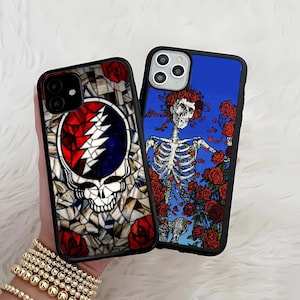 May include: Two phone cases with black borders. One features a stained-glass design of a skull with a lightning bolt and red roses. The other has a skeleton with red roses on a blue background. Both cases have a glossy finish.