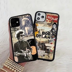 May include: Two black phone cases featuring Bob Dylan imagery. One case has a collage design with text and a photo of Dylan playing guitar. The other case has a vintage poster design with Dylan's name and image.