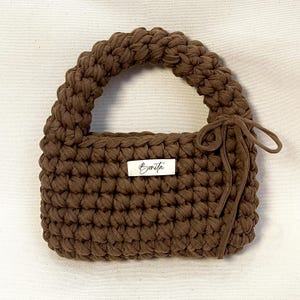 Pode incluir: Bolsa de crochê marrom com corpo retangular e alça arredondada. A bolsa apresenta uma pequena etiqueta com a palavra "Bonita" em escrita cursiva. Um detalhe de laço correspondente adiciona um elemento decorativo na lateral.
