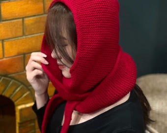 May include: A vibrant red knitted hood and scarf combination. The hood covers the head and neck, with long scarf ends. The thick knit texture is visible. The garment is worn over a black top.