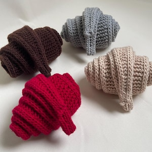 May include: Four knitted croissants in shades of brown, gray, red, and beige. The handmade pastries are arranged on a white surface. The croissants are made with a textured stitch, giving them a realistic appearance.