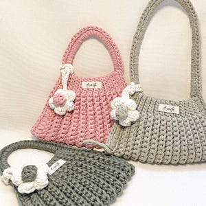 May include: Three handmade crochet handbags in pink, gray, and green. Each bag features a flower accent and a label that reads "Barita". The bags have braided handles and a textured stitch pattern.
