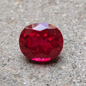 May include: A vibrant, deep red gemstone with a faceted oval cut. The stone features a highly reflective surface with numerous geometric facets that catch the light, creating a brilliant, sparkling appearance across its polished, translucent body.