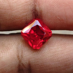 May include: A vibrant, square-cut gemstone with a deep red colour and multiple facets that reflect light. The stone features a sharp, geometric shape with pointed corners and a polished, translucent surface that displays internal light refraction.