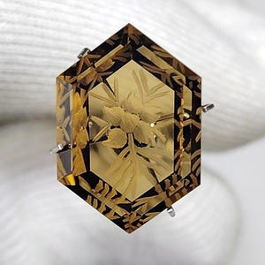 May include: A hexagonal, amber-colored gemstone featuring an intricate, carved floral design on its surface. The gem has a faceted border with dark, geometric patterns and is held by small, silver-toned metal prongs on its sides.