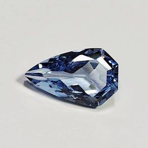 May include: A pear-shaped faceted gemstone with a translucent light blue color. The stone features a pointed end and a wider, rounded end with multiple geometric facets that reflect light, creating a brilliant and clear appearance throughout.
