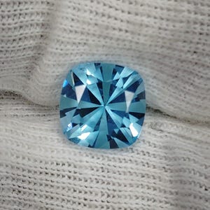 May include: A cushion-cut gemstone with a vibrant light blue color and a faceted surface that creates a starburst pattern of light reflections. The stone has rounded corners and a square-like shape, displaying high clarity and brilliance.