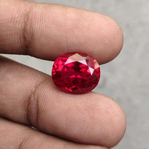 May include: A faceted, oval-shaped, deep red gemstone. The gem is held between two fingers, showcasing its brilliant cut and vibrant color. The stone's facets reflect light, creating a sparkling effect.