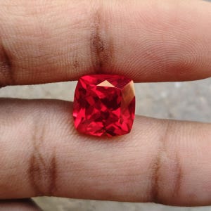 Könnte beinhalten: Ein facettierter, kissenförmiger Edelstein in leuchtendem Rot. Der Stein wird zwischen Fingern gehalten, was seine Brillanz und Klarheit zeigt. Die quadratische Form mit abgerundeten Ecken reflektiert das Licht.