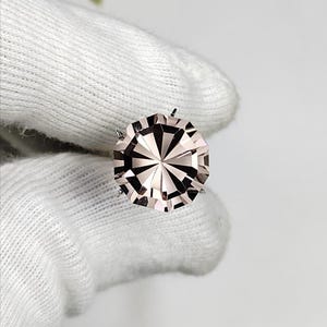 Puede incluir: Una gema redonda y facetada de color rosa pálido. La gema está sujeta por pequeñas garras metálicas. El corte de la gema crea un patrón de estrella. El fondo es blanco. Diámetro aproximado de 1,5 cm.