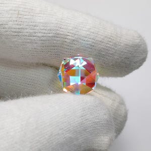 Könnte beinhalten: Eine facettierte, irisierende Kristallkugel mit geometrischem Design. Der Kristall reflektiert ein Farbspektrum, darunter Rosa, Orange, Blau und Grün. Die Kugel wird in einem weißen Handschuh gehalten, was ihre Brillanz hervorhebt.