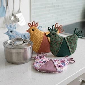 May include: Four fabric chicken-shaped pot holders in blue floral, yellow polka dot, green cherry print, and purple floral patterns, arranged around a stainless steel pot with a lid. Each chicken features a small black button eye and fabric loops.