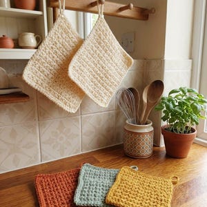 Könnte beinhalten: Fünf quadratische Häkel-Topflappen mit strukturiertem Waffelmuster. Zwei cremefarbene Topflappen hängen an einem Holzständer, während drei weitere in Rostorange, Salbeigrün und Senfgelb auf einer Holzoberfläche angeordnet sind.
