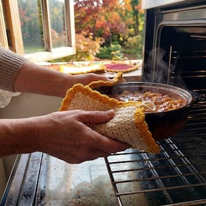 May include: A cream-coloured crochet potholder with a bright yellow scalloped border. The potholder is being used to hold a dark brown ceramic baking dish filled with a steaming, cooked meal as it is being removed from a metal oven rack.