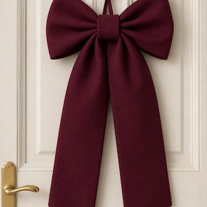 May include: A large burgundy bow hangs on a white door. The bow is made of a thick fabric and has long, flowing tails. A gold-colored door handle and keyhole are visible on the door.