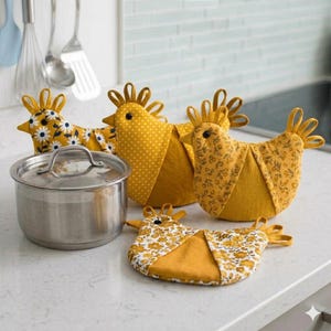 Puede incluir: Tres fundas para tetera de tela con forma de gallina en tonos amarillo, mostaza y blanco, con estampados florales y de lunares, junto con un agarrador de tela a juego con forma de gallina y una olla de acero inoxidable con tapa.