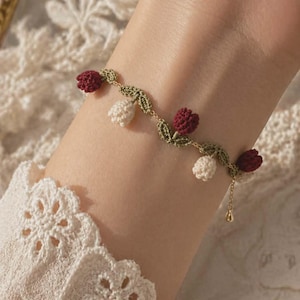 May include: A delicate, handmade bracelet featuring a floral design. The bracelet has a gold chain with crocheted red and white flowers and green leaves. The bracelet is approximately 7 inches (17.78 cm) in length.