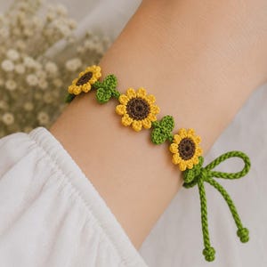 May include: Crocheted bracelet featuring yellow and brown sunflowers with green leaves. The bracelet is tied with a green string and bow. The bracelet is a charming accessory.