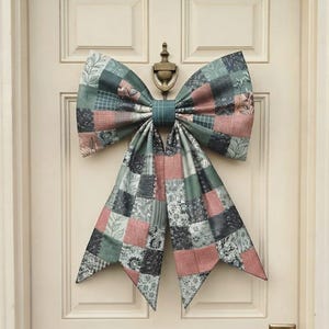 May include: A large decorative bow made of fabric with a patchwork design featuring squares in shades of sage green, dusty pink, and charcoal gray. The fabric displays various patterns including floral motifs, leaves, and grid lines, with two long tails hanging down from the center knot.