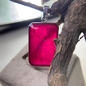 May include: A rectangular pendant with a vibrant magenta gemstone. The gemstone is set in a silver-colored metal frame with a simple bail. The pendant is displayed against a neutral background, highlighting its color and shape.