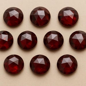 Puede incluir: Diez gemas redondas de color rojo oscuro con superficies facetadas se disponen sobre un fondo beige claro. Cada gema tallada crea múltiples superficies reflectantes, produciendo un efecto brillante. Las gemas están espaciadas uniformemente.