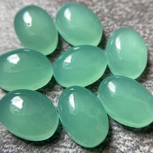 May include: Eight oval-shaped, polished gemstones in a soft, seafoam green hue. The stones have a smooth, glossy surface, reflecting light. The gemstones are arranged in a scattered pattern, showcasing their individual shapes and color.